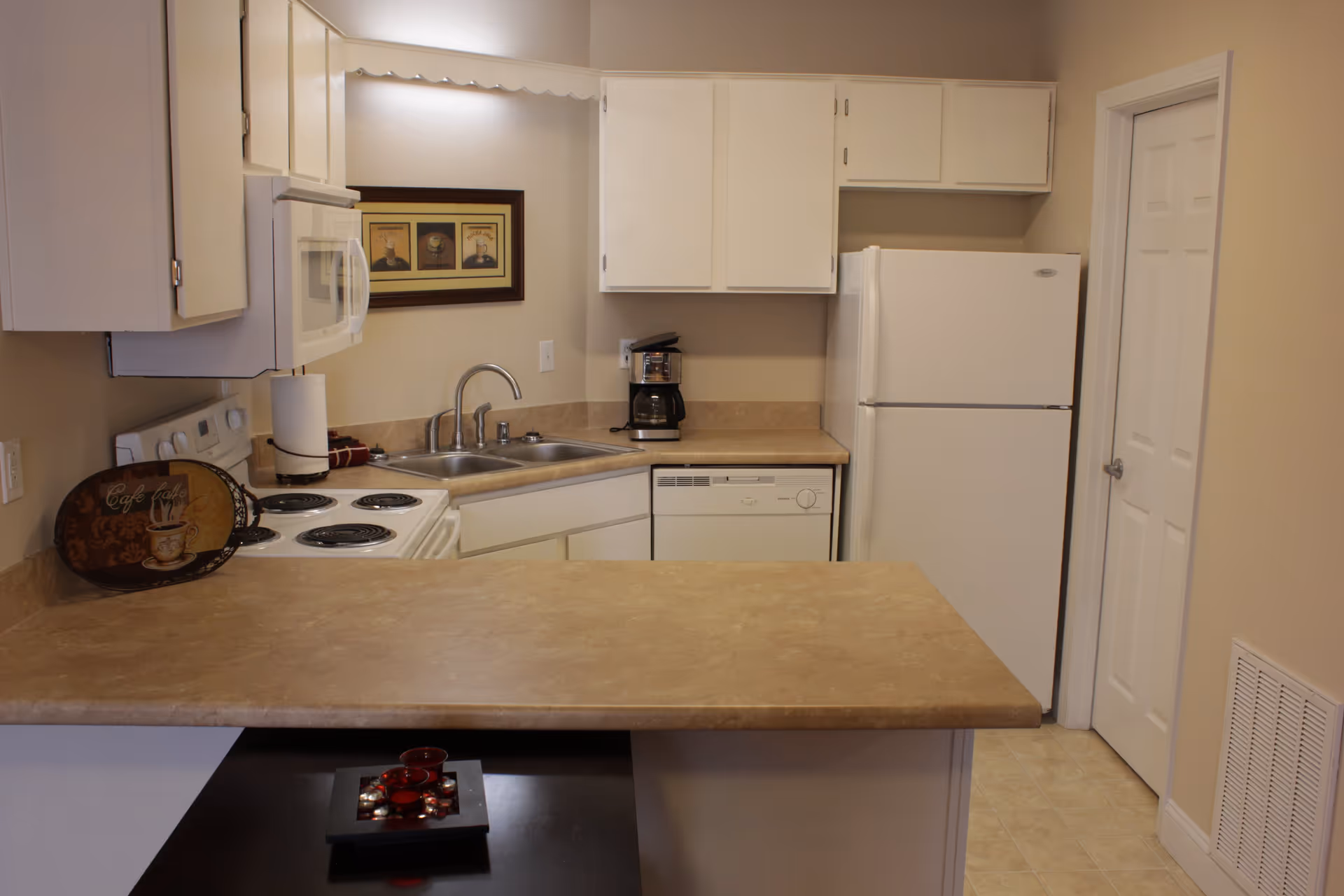 A compact kitchen with beige countertops and white cabinets. The kitchen features a white refrigerator, dishwasher, microwave, electric stove, double sink, and a coffee maker. There is a framed picture on the wall above the sink and a decorative tray on the counter.