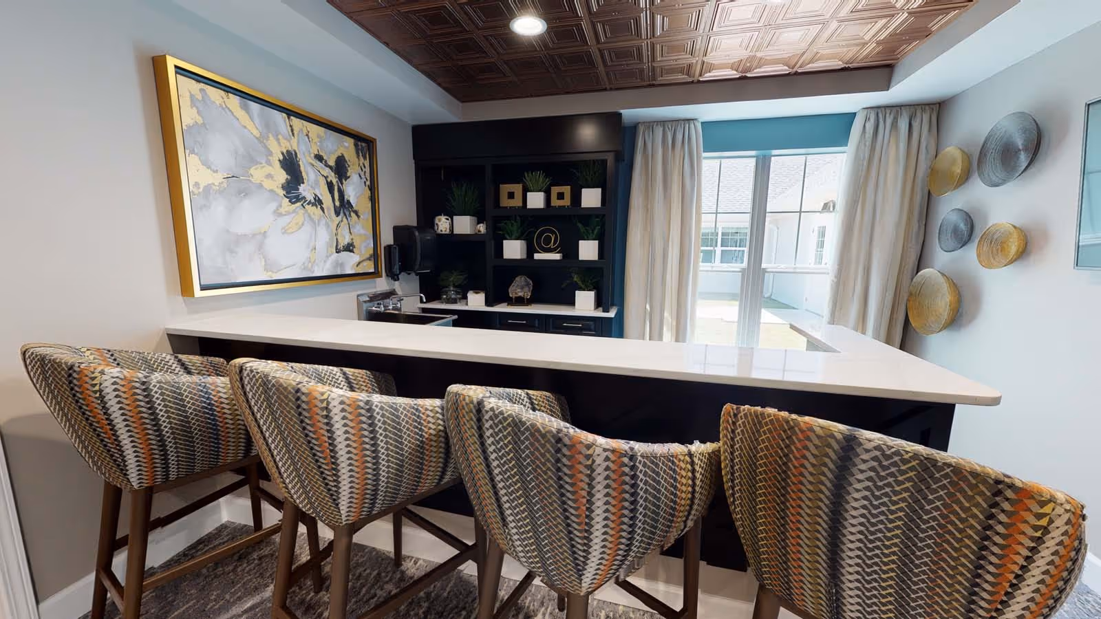 Interior view of a modern lounge area with a white countertop bar and four patterned upholstered bar stools. Behind the bar is a black shelving unit with decorative items and plants. A large abstract painting with gold, white, and gray tones hangs on the wall to the left. The ceiling features a decorative copper-colored tile design. A sliding glass door with beige curtains lets in natural light, and decorative round wall hangings are displayed on the right wall.
