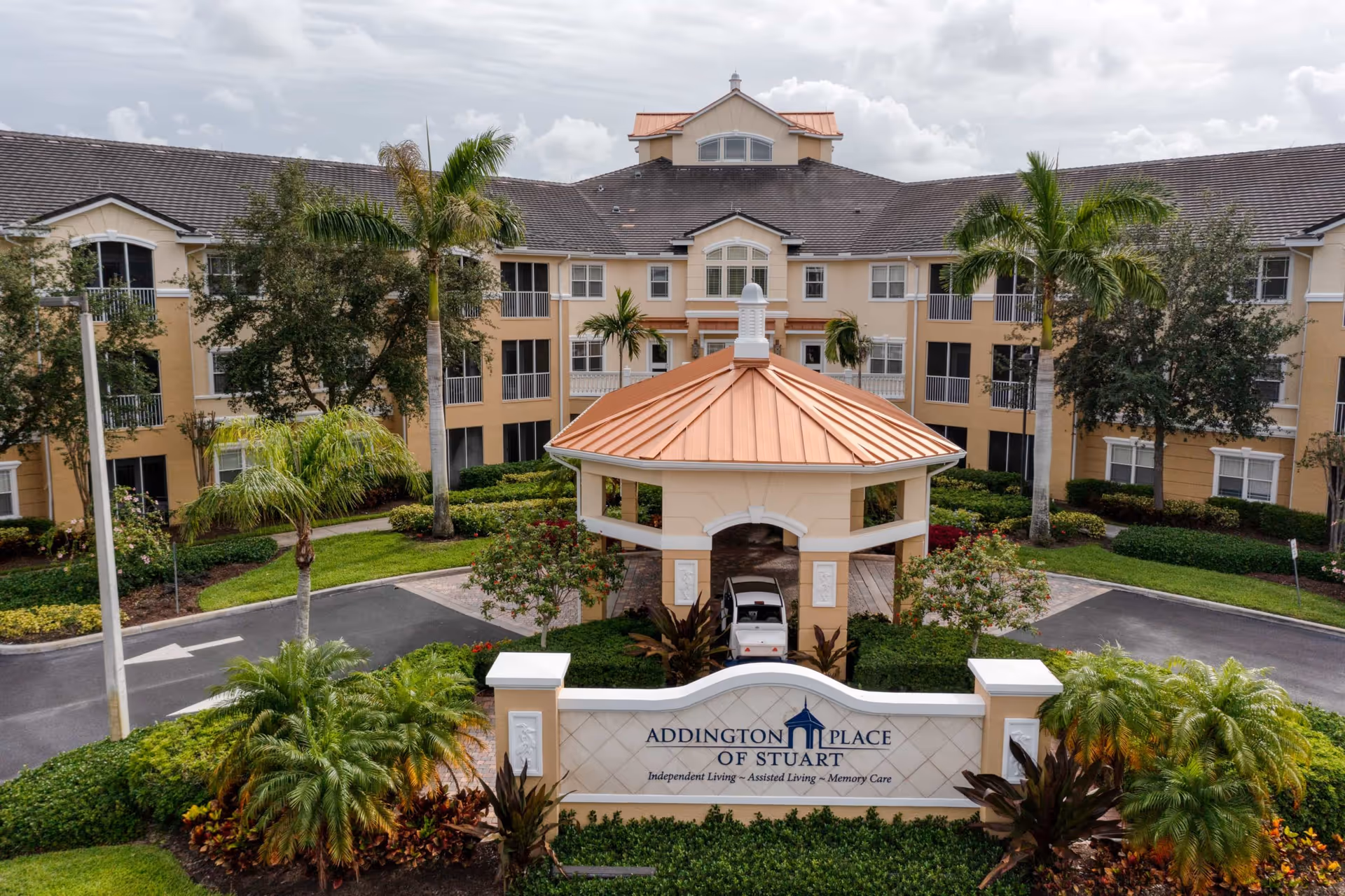 Exterior view of Addington Place of Stuart, a senior living facility with a three-story beige building, palm trees, and a covered entrance with a copper-colored roof. The facility sign is visible in front, surrounded by landscaped greenery.