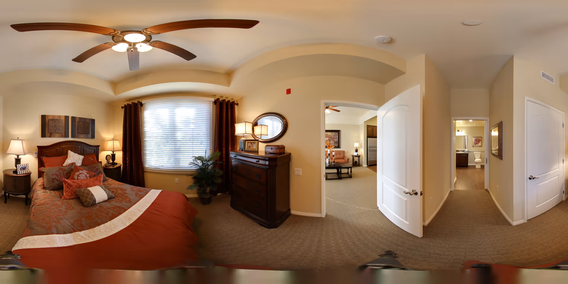 A warmly lit bedroom with a large bed adorned with multiple pillows and a patterned bedspread. The room features a ceiling fan with lights, two bedside tables with lamps, a window with closed blinds and dark curtains, a wooden dresser with a lamp and mirror above it, and an open door leading to a living area and a hallway with a bathroom visible at the end.