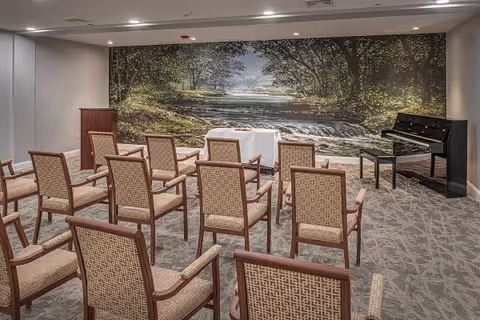 A small meeting or event room with rows of wooden chairs facing a table covered with a white cloth. A black piano with a bench is positioned to the right side of the room. The back wall features a large mural of a serene forest scene with a river running through it. The room has neutral-colored walls and carpeted flooring.