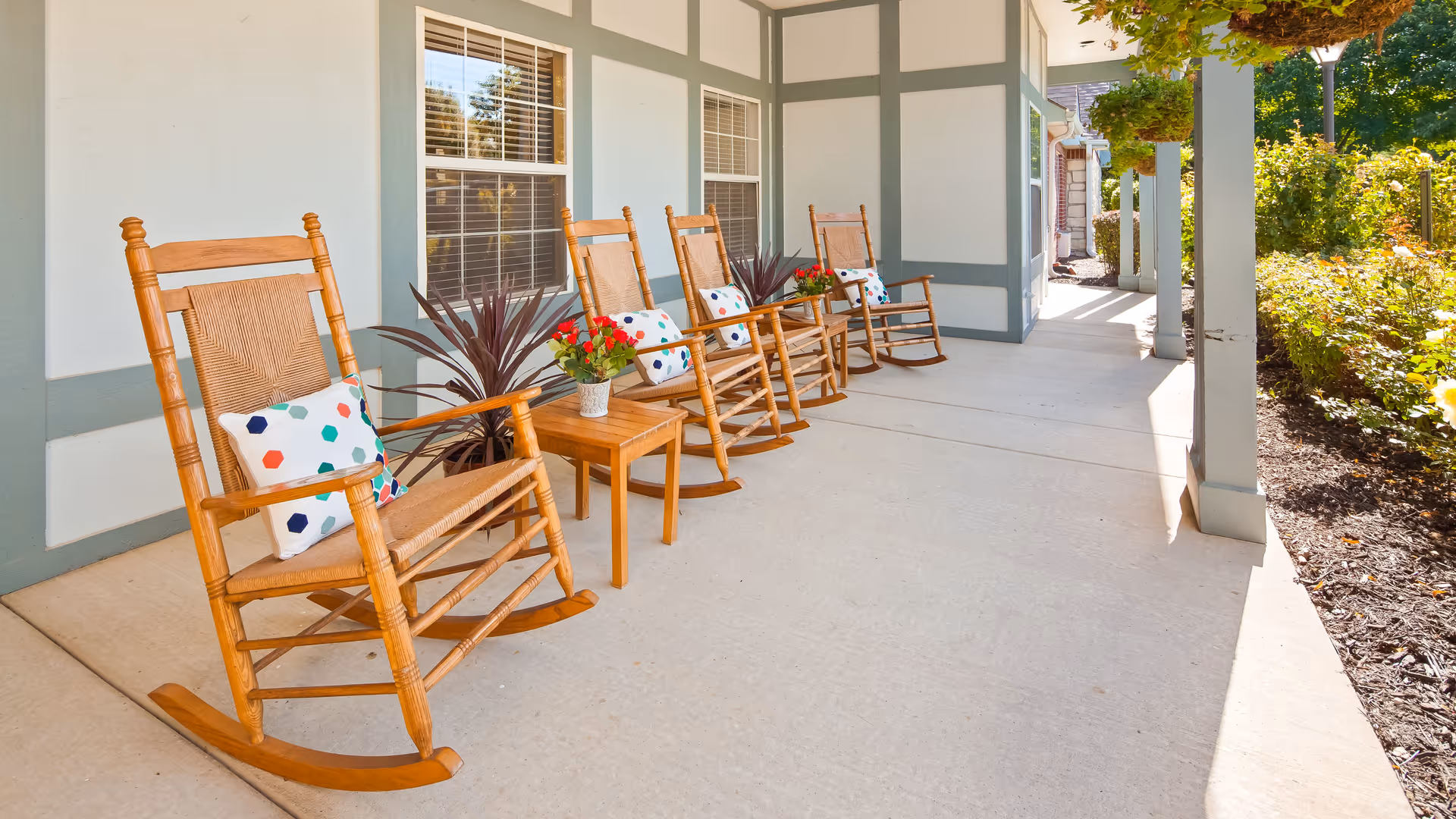A covered outdoor porch area with four wooden rocking chairs, each with a colorful polka dot cushion. There are small wooden tables between the chairs with potted plants on them. The porch is adjacent to a garden with green bushes and flowers, and the building has light-colored walls with green trim.