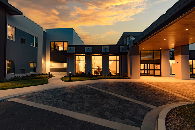 Illuminated entrance and porte-cochère of a modern senior living building at sunset.