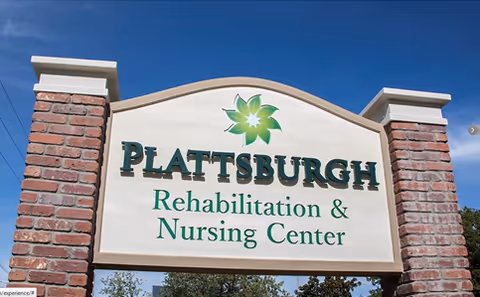 Entrance sign reading "Plattsburgh Rehabilitation & Nursing Center" mounted between brick pillars against a blue sky.