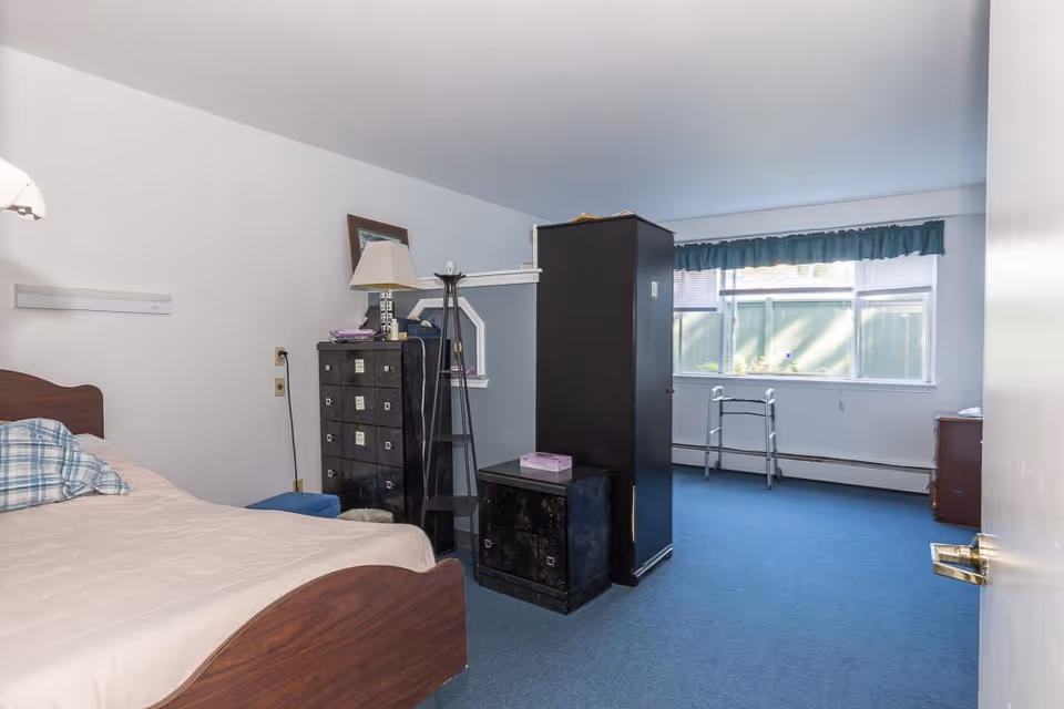 A senior living facility bedroom with a wooden bed featuring a beige bedspread and a plaid pillow. The room has blue carpet flooring, a black dresser with a lamp and mirror on top, a tall black wardrobe, and a window with blue curtains letting in natural light. A walker is positioned near the window.
