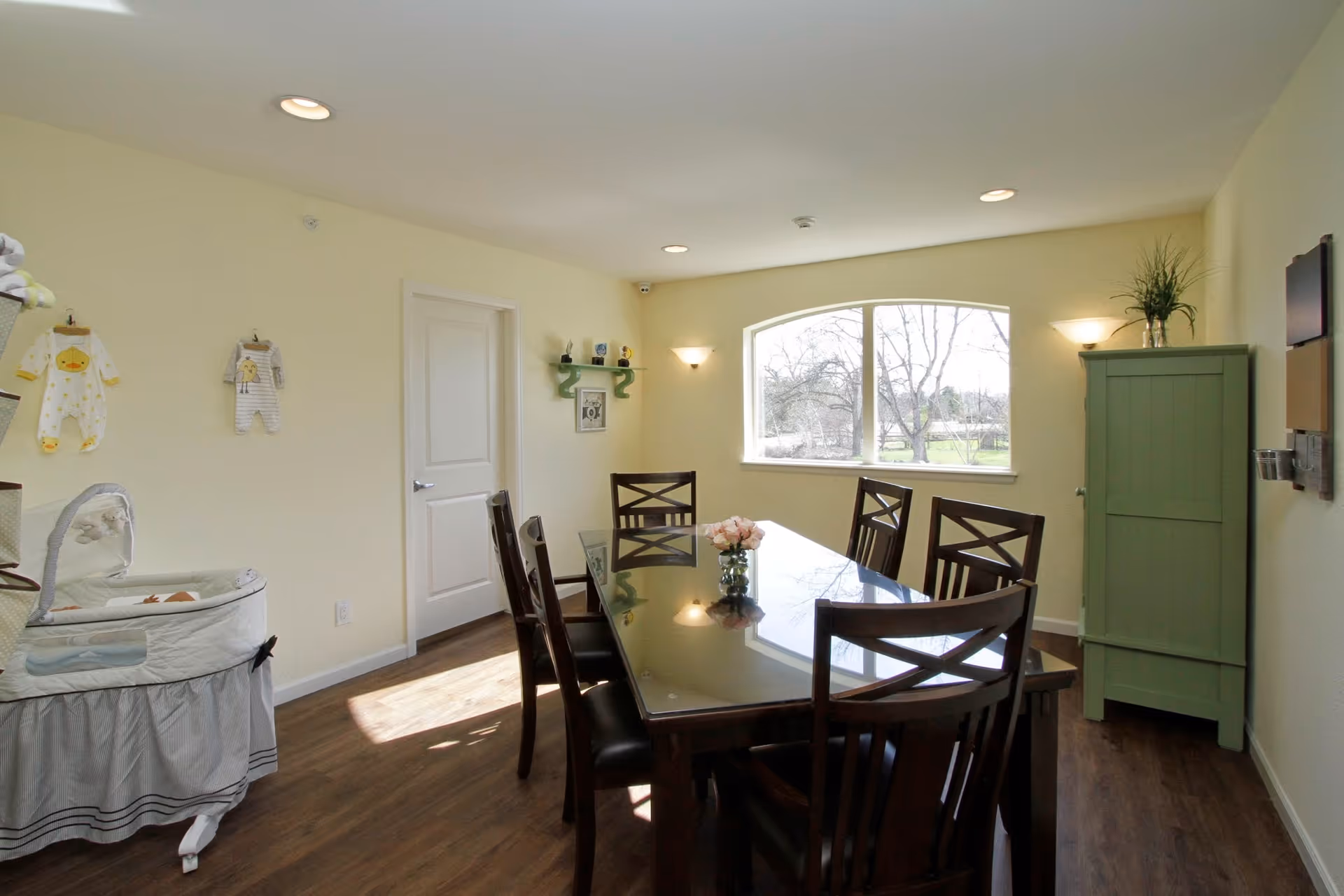 A well-lit dining room with a large rectangular glass-top table surrounded by six wooden chairs. There is a green cabinet in the corner with a plant on top, a window showing trees outside, and baby clothes hanging on the left wall near a bassinet. The room has light yellow walls and wooden flooring.