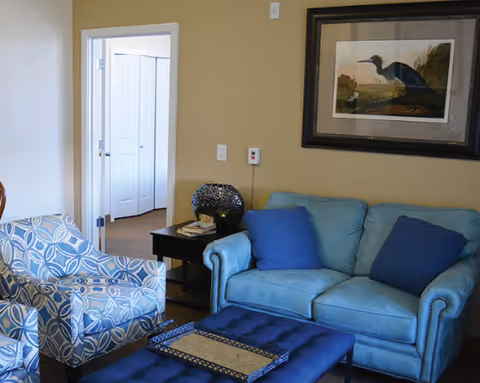 A cozy living room featuring a light blue loveseat with two dark blue pillows, a patterned blue and white armchair, and a dark blue tufted ottoman with a decorative tray on top. A small black side table with books and a decorative lamp is placed next to the loveseat. A framed picture of a bird hangs on the beige wall above the loveseat. An open doorway reveals a glimpse of another room with white double doors.