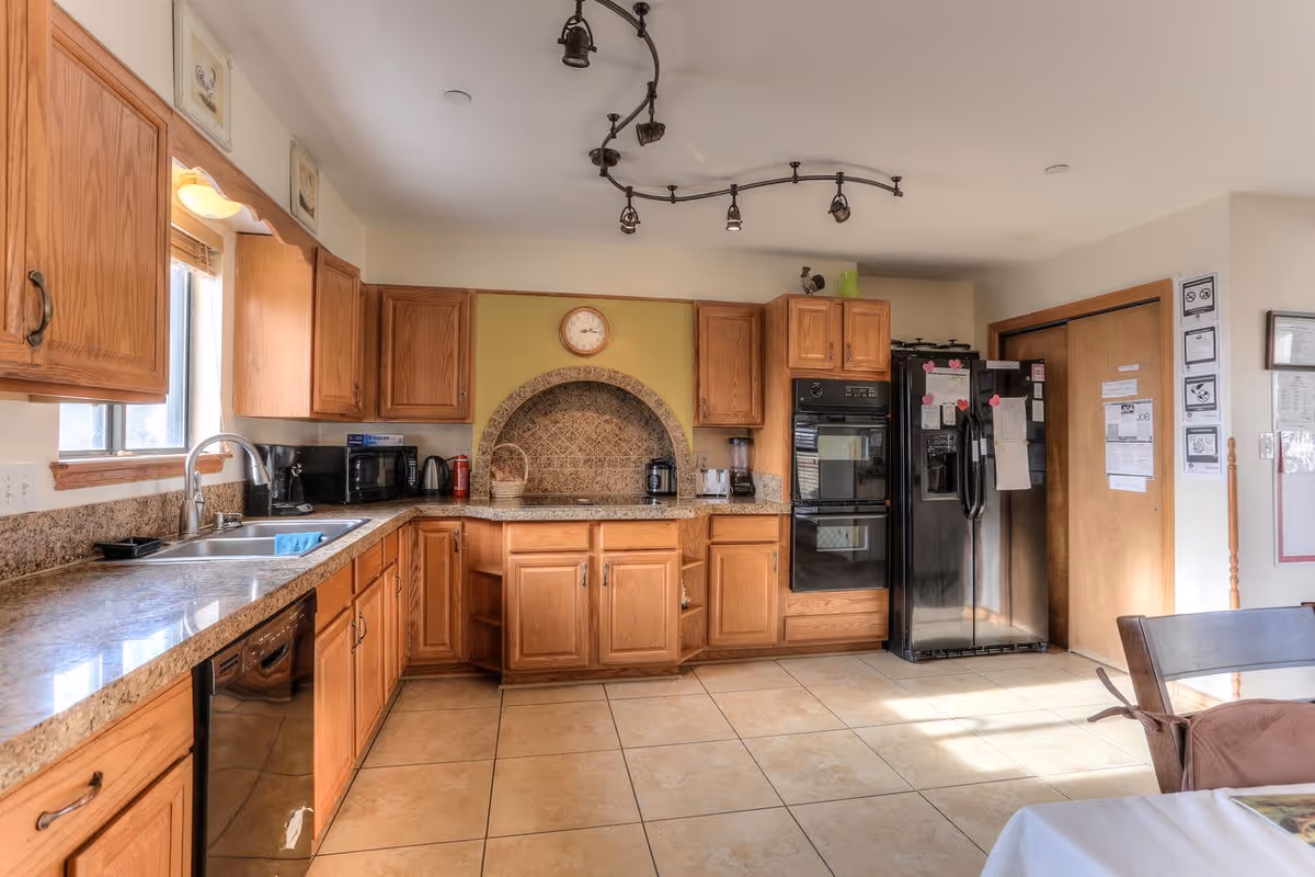 A spacious kitchen with wooden cabinets, granite countertops, and tiled floor. The kitchen features a double sink under a window, a dishwasher, a microwave, a coffee maker, a double oven, and a black refrigerator. There is a curved track light fixture on the ceiling and various papers and notes attached to the refrigerator and the wall near a wooden door.