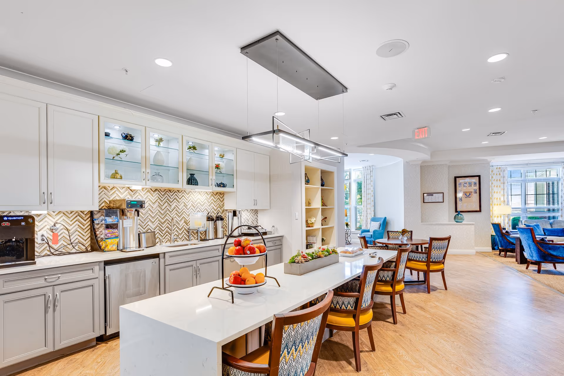 Bright communal kitchen and dining area with a long island, chairs, cabinets, and tables in a senior living facility.
