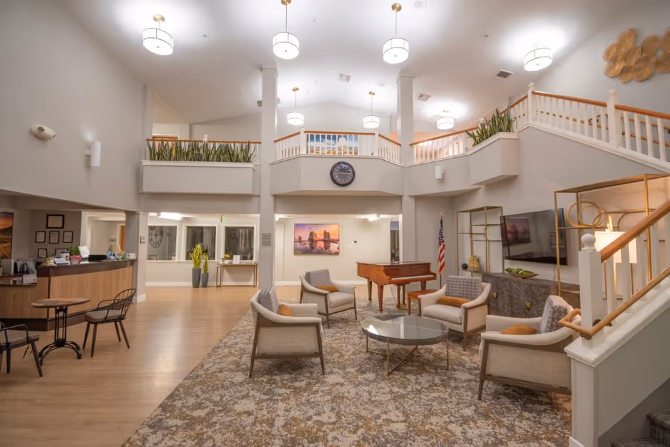 A spacious and well-lit senior living facility common area with a high ceiling and upper balcony. The room features a seating area with six cushioned chairs arranged around a glass coffee table on a patterned carpet. A grand piano is positioned near the back wall, which also displays a large wall clock and a scenic framed picture. To the left, there is a reception desk with chairs and a small round table with two chairs. The area is decorated with plants, modern light fixtures, and an American flag near the piano. A staircase with wooden handrails leads to the upper level.