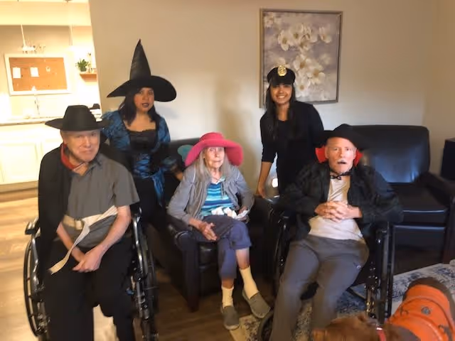A group of five people in a living room setting, three elderly individuals seated in chairs and wheelchairs wearing hats and costumes, accompanied by two younger adults also dressed in costumes, including a witch hat and a police hat. The room has a painting on the wall and a kitchen area visible in the background.