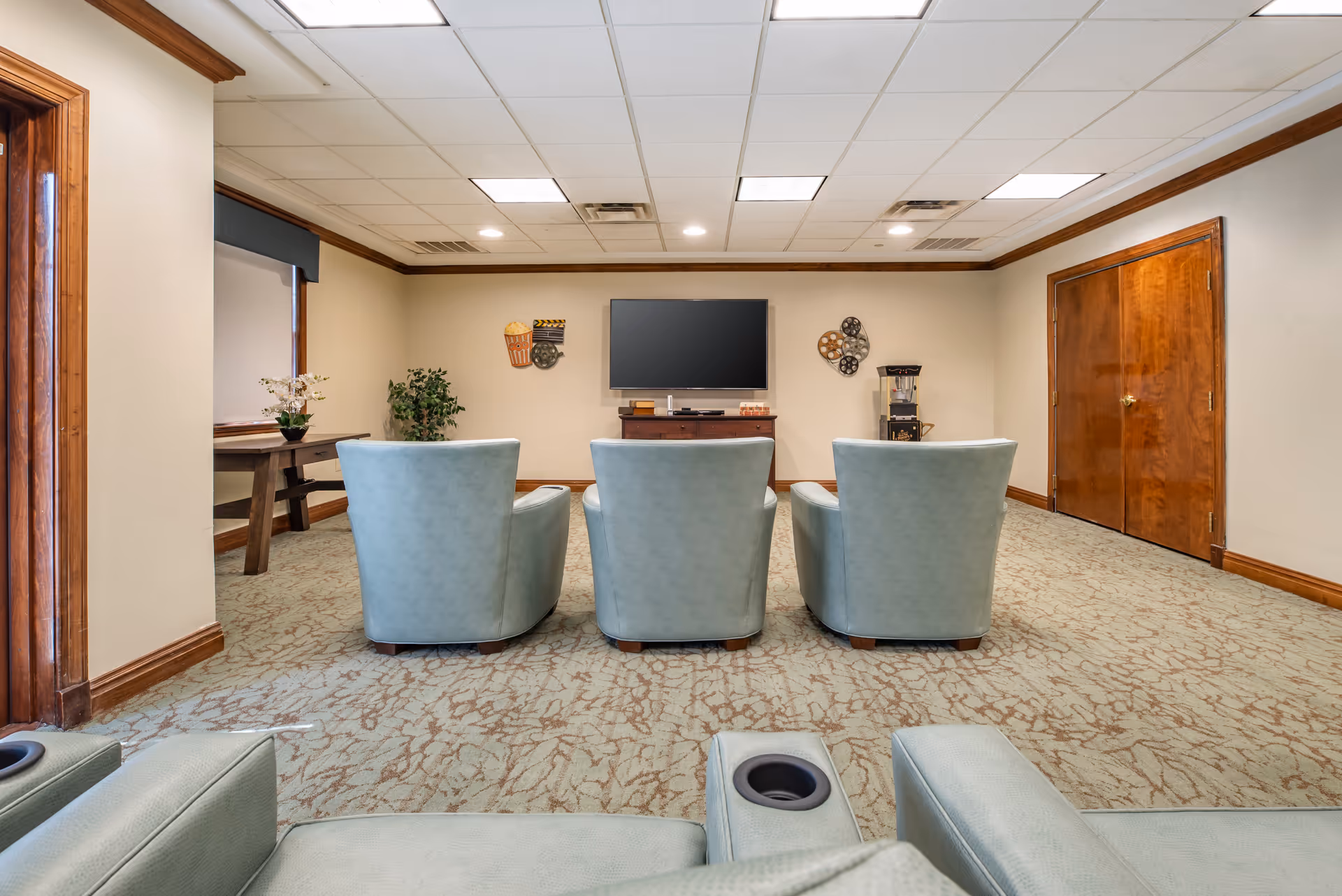 A cozy media room with three light blue armchairs facing a flat-screen TV mounted on a beige wall. The room has a patterned carpet, wooden trim, and a popcorn machine on the right side. There are decorative movie-themed wall hangings and a wooden table with a plant on the left side.