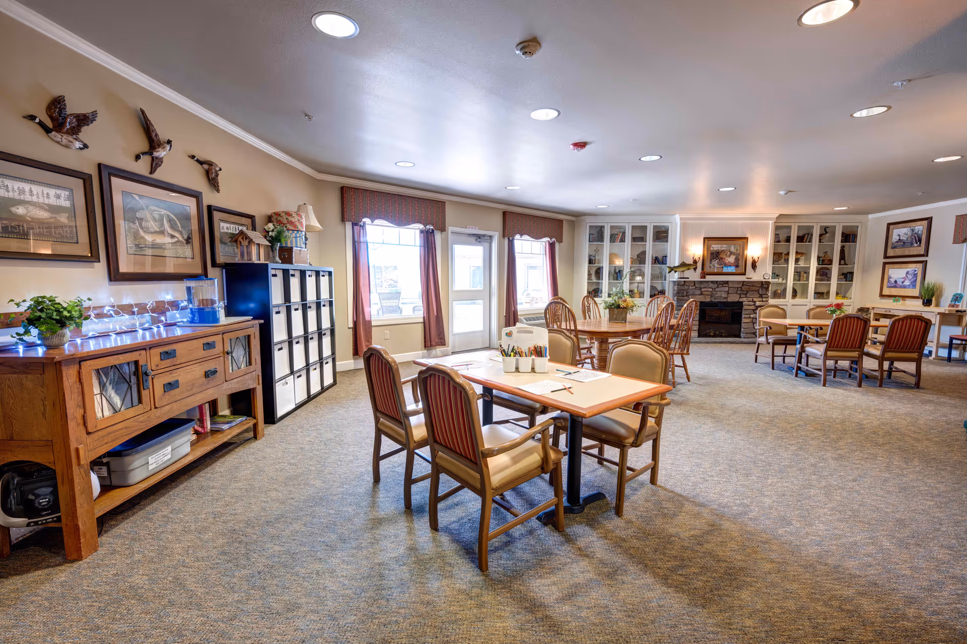 A spacious, well-lit common area in a senior living facility with multiple tables and chairs arranged for activities or dining. The room features a carpeted floor, a stone fireplace with built-in shelves on either side, framed artwork and decorative ducks mounted on the wall, and windows with curtains allowing natural light to enter.