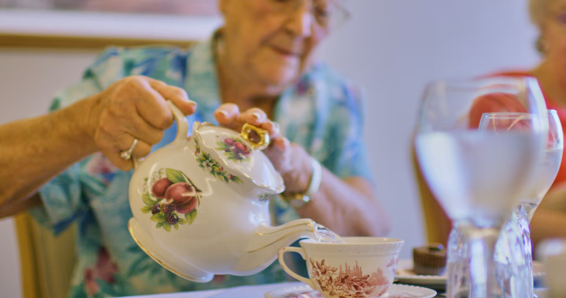 An elderly woman wearing a blue floral shirt is pouring tea from a white teapot decorated with fruit designs into a matching teacup on a table set with glasses of water and a small dessert.