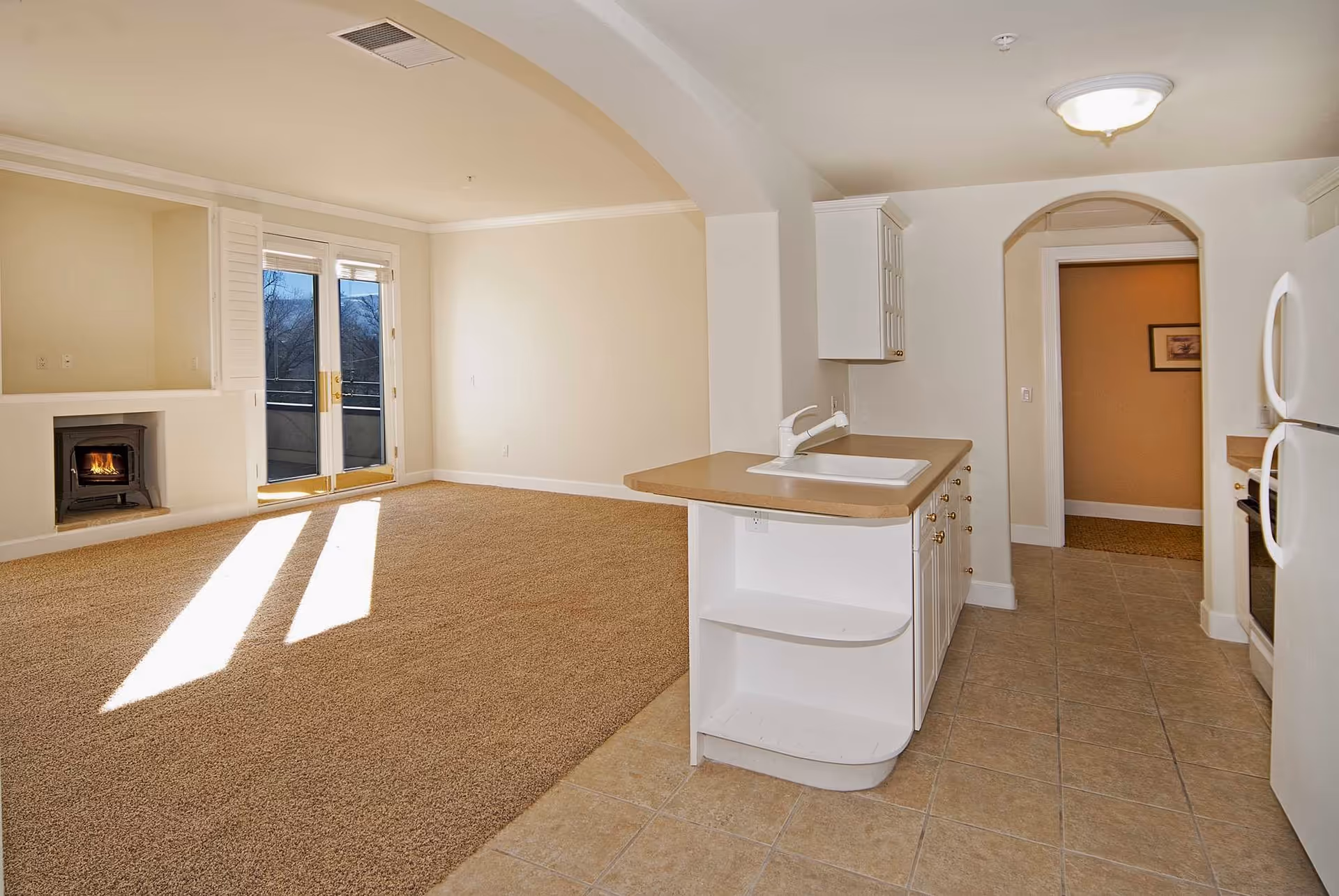 An empty open-concept living area and kitchen in a senior living facility. The living area has beige carpet, a small fireplace with a fire burning, and double glass doors leading to an outdoor balcony. The kitchen has tiled flooring, white cabinets, a countertop with a sink, and white appliances including a refrigerator and oven. The walls are painted light beige, and there is an arched doorway leading to another room.