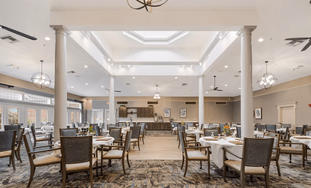 A spacious and well-lit dining room with multiple tables covered with white tablecloths and set with napkins and small flower arrangements. The room features tall white columns, large windows allowing natural light, modern chandeliers, ceiling fans, and a serving area at the back with cabinets and coffee machines.