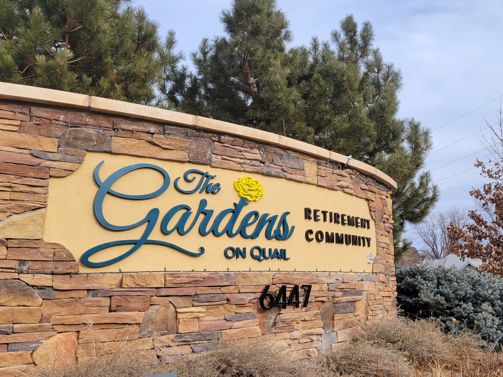 Stone and stucco sign for The Gardens on Quail Retirement Community with the address number 6447, surrounded by trees and shrubs under a partly cloudy sky.