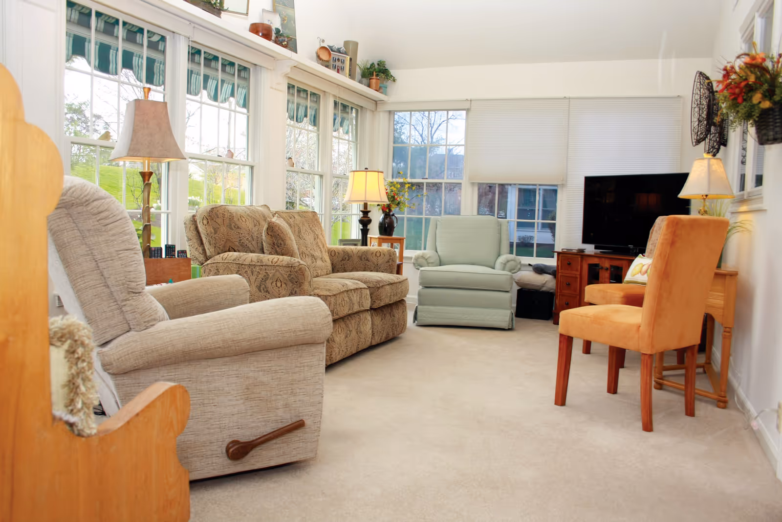 Sunlit living room with multiple upholstered chairs, lamps, a TV, and large windows.