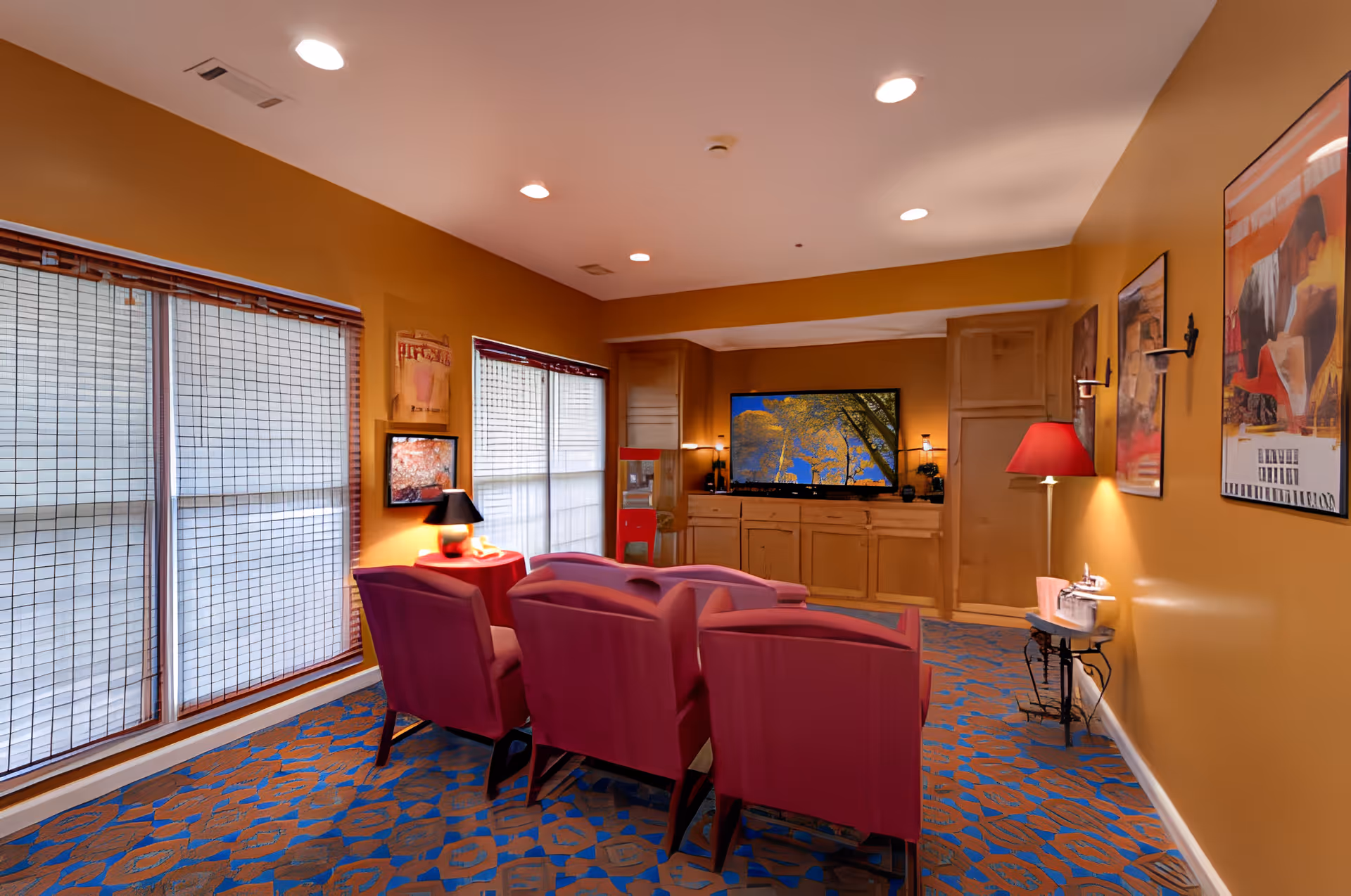 A cozy living room with mustard yellow walls and a patterned blue and orange carpet. There are six red upholstered chairs arranged in two rows facing a large flat-screen TV mounted on a wooden cabinet. The room is softly lit with recessed ceiling lights, a table lamp on a small round table, and a floor lamp with a red shade. Large windows with blinds cover one wall, and framed artwork decorates the opposite wall.