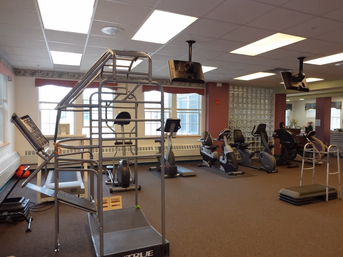 Exercise room with cardio machines, strength-training frame, stacked step platforms and ceiling-mounted TVs in a senior living facility.