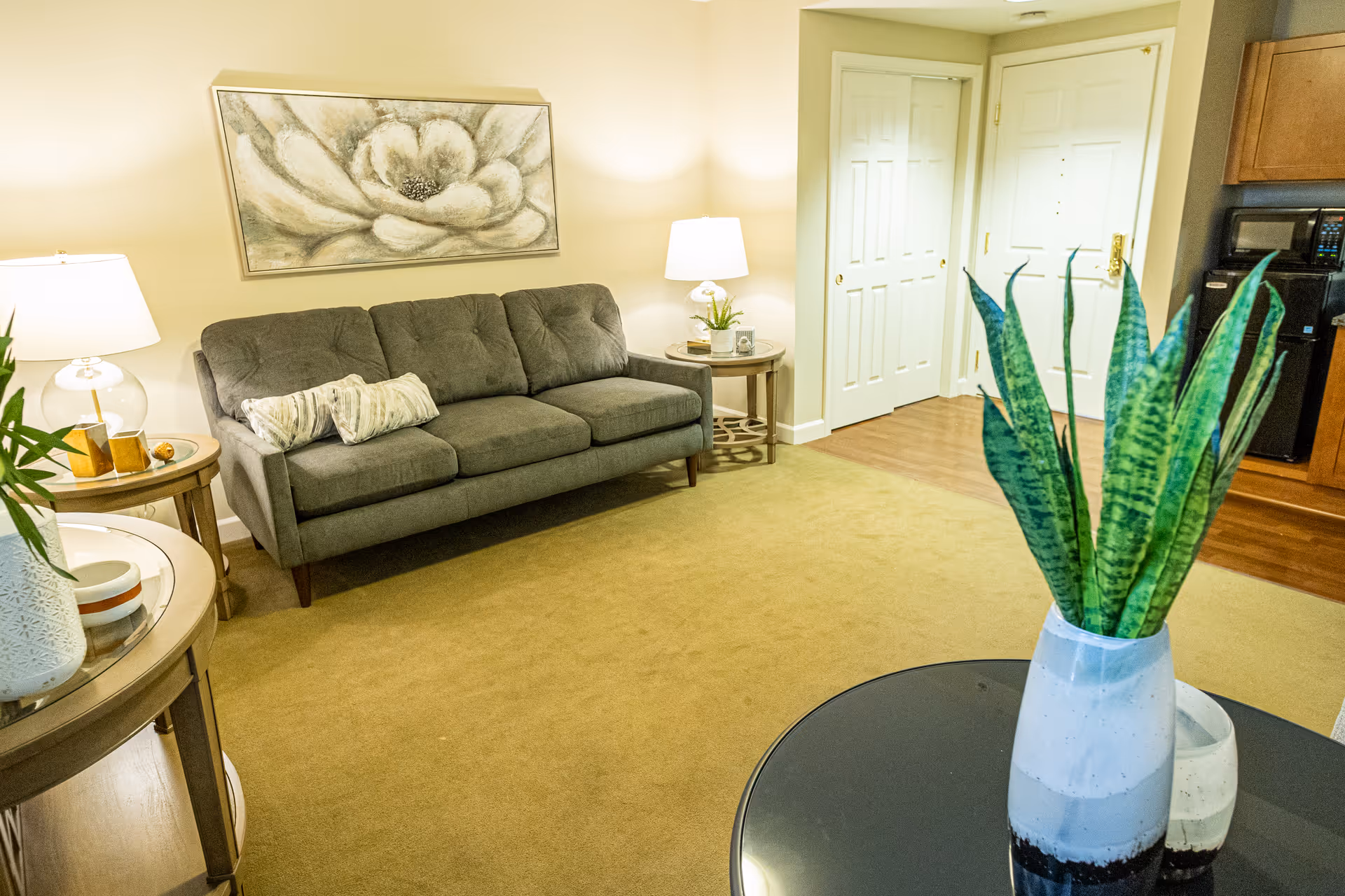 A cozy living room with a gray three-seater sofa adorned with two striped pillows, flanked by two round wooden side tables each holding a lamp. A large floral painting hangs above the sofa. The room has beige walls and carpet, with a white door and closet in the background. In the foreground, there is a round black table with a tall green plant in a white and black vase.