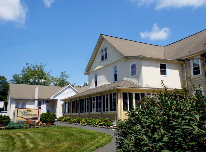 Front exterior of the Welsh Mountain Home building with a screened porch, landscaped lawn, and entrance sign under a blue sky.