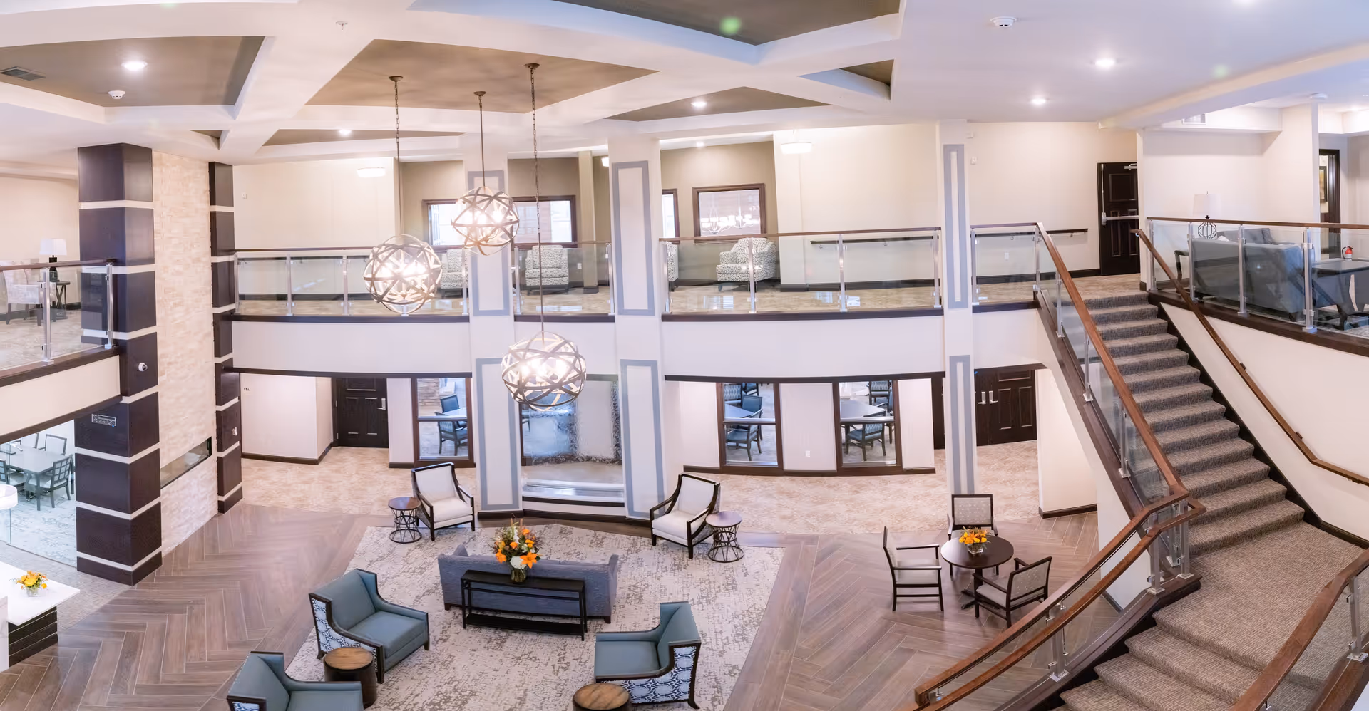 Spacious and well-lit senior living facility common area with modern furniture including armchairs, sofas, and small tables. The room features a high ceiling with three spherical pendant lights hanging down. There is a staircase with glass and wood railing leading to an upper level with additional seating areas. The walls are painted in neutral tones with decorative panels, and the floor has a combination of carpet and wood-like tiles.