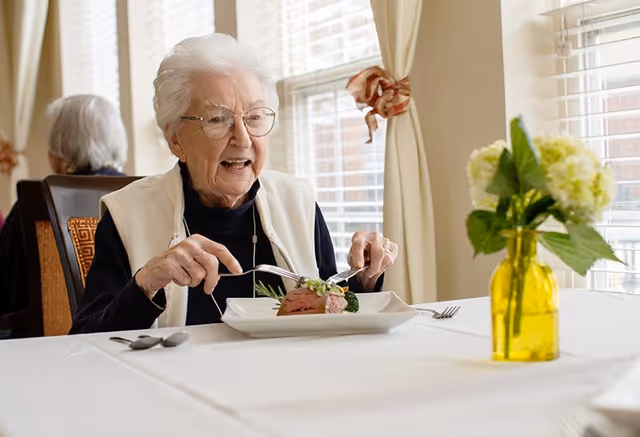 An elderly woman with white hair and glasses is sitting at a dining table in a well-lit room, cutting into a plated meal. There is a yellow vase with green and white flowers on the table, and another elderly person is visible in the background near a window with white blinds and cream-colored curtains.