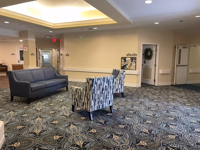 A spacious interior common area with patterned carpet, a gray sofa, and two patterned armchairs. The walls are light beige with white trim, and there is a hallway extending into the distance. A bulletin board and a door decorated with a green wreath are visible on the right side.