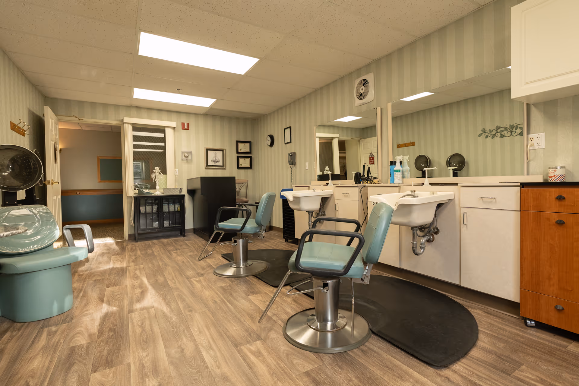 Interior view of a senior living facility's hair salon with two teal salon chairs in front of sinks and mirrors, a hair dryer chair on the left, wood-patterned flooring, and light green striped wallpaper.