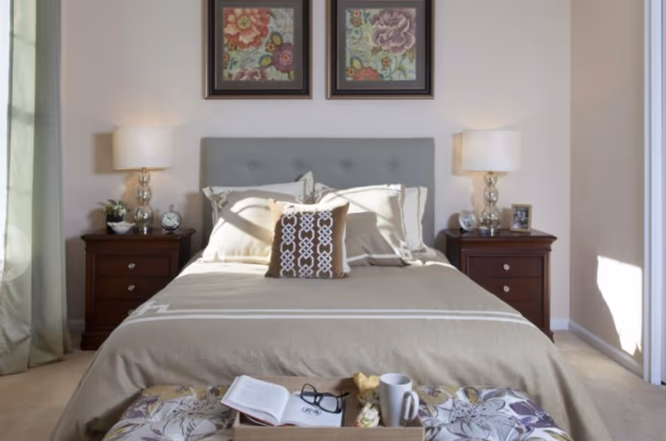 A neatly made bed with beige bedding and a decorative pillow in the center, flanked by two dark wooden nightstands each with a lamp, clock, and small decor items. Above the bed are two framed floral paintings. At the foot of the bed is a cushioned bench with an open book, glasses, a mug, and a small wrapped item on it. Light streams in from a window on the right side of the room.
