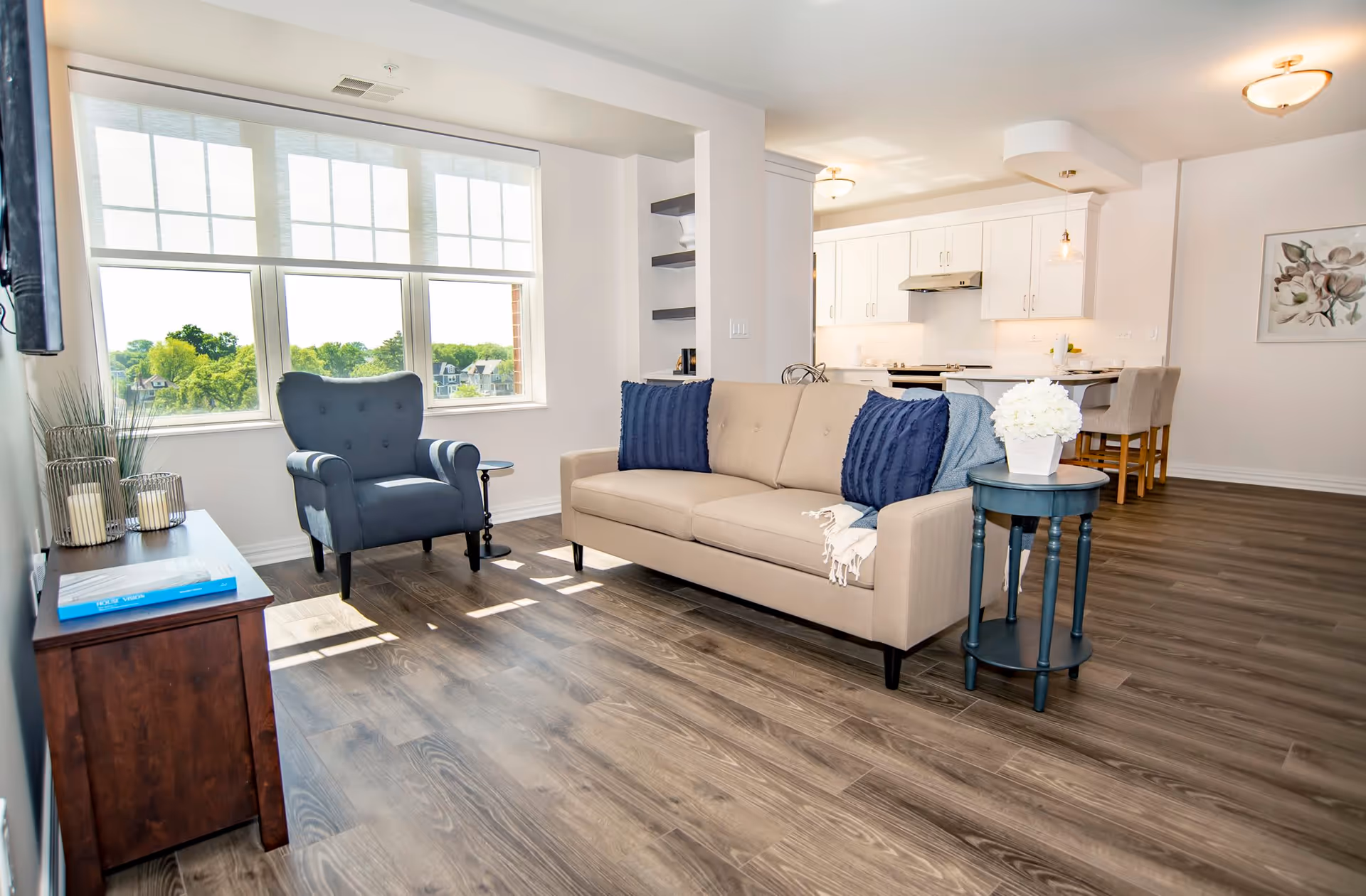 Bright open living room with a beige sofa, blue armchair, round side table and large windows overlooking greenery, with a kitchen visible in the background.