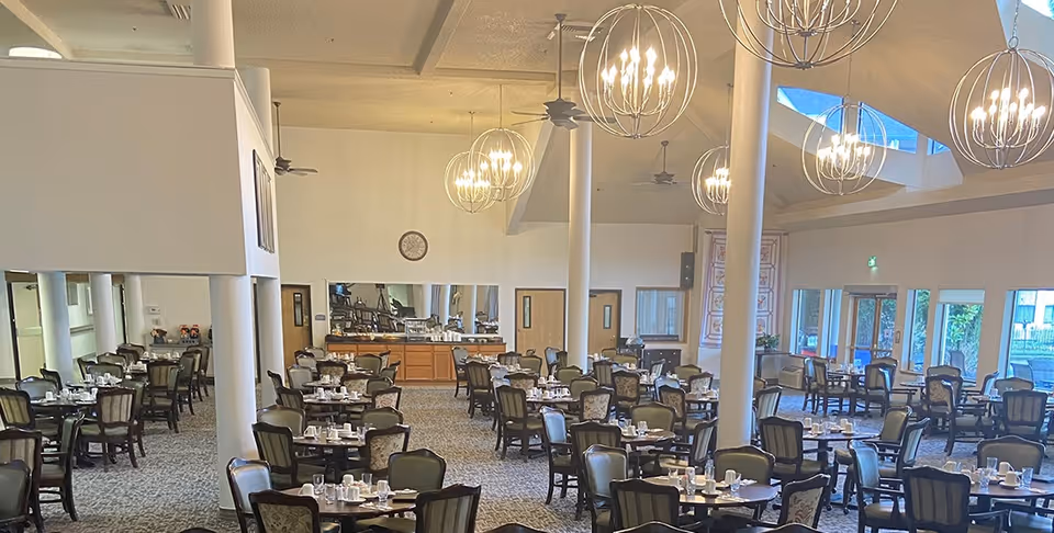 Spacious dining room with multiple round tables set with cups, plates, and silverware. The room features high ceilings with modern spherical chandeliers, ceiling fans, and large windows allowing natural light to enter. There are several chairs around each table and a buffet area with mirrors on the far wall.