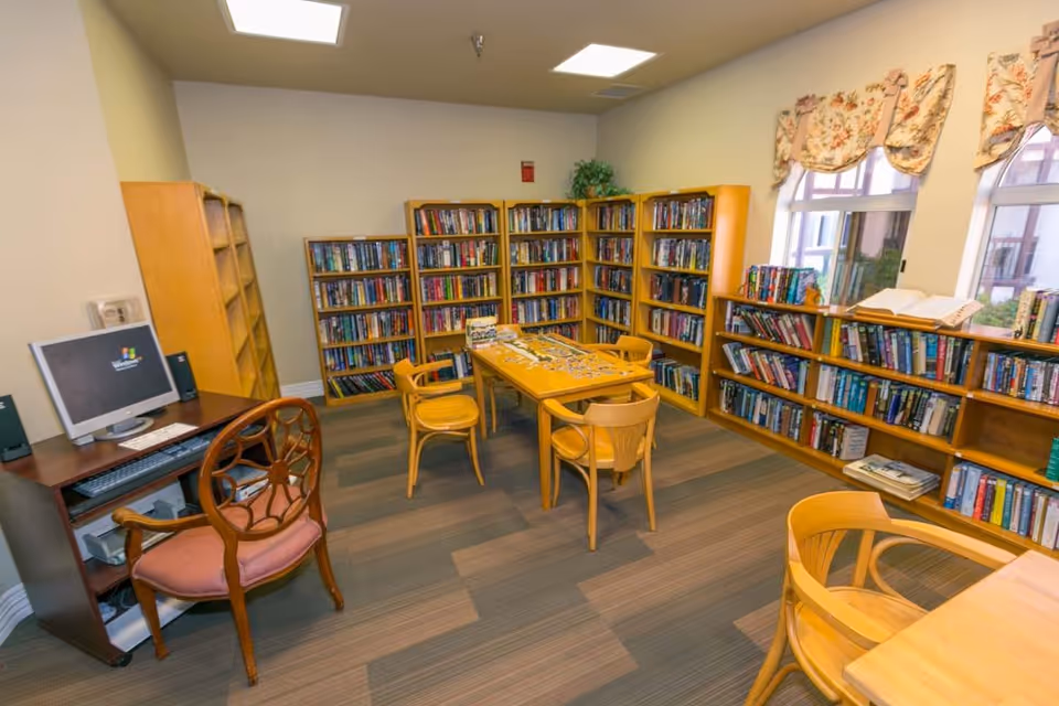 A cozy library room with wooden bookshelves filled with books along the walls. There is a wooden table in the center with four wooden chairs around it. A computer desk with a monitor and a wooden chair with a pink cushion is positioned on the left side of the room. The room has carpeted flooring and windows with floral valances letting in natural light.