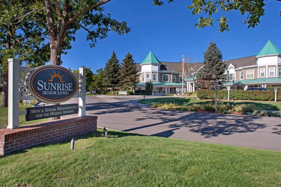 Exterior view of Sunrise Senior Living facility with a sign in the foreground that reads 'Sunrise Senior Living with a special neighborhood for the memory impaired.' The building has green roofs, multiple windows, and is surrounded by trees and landscaped greenery under a clear blue sky.