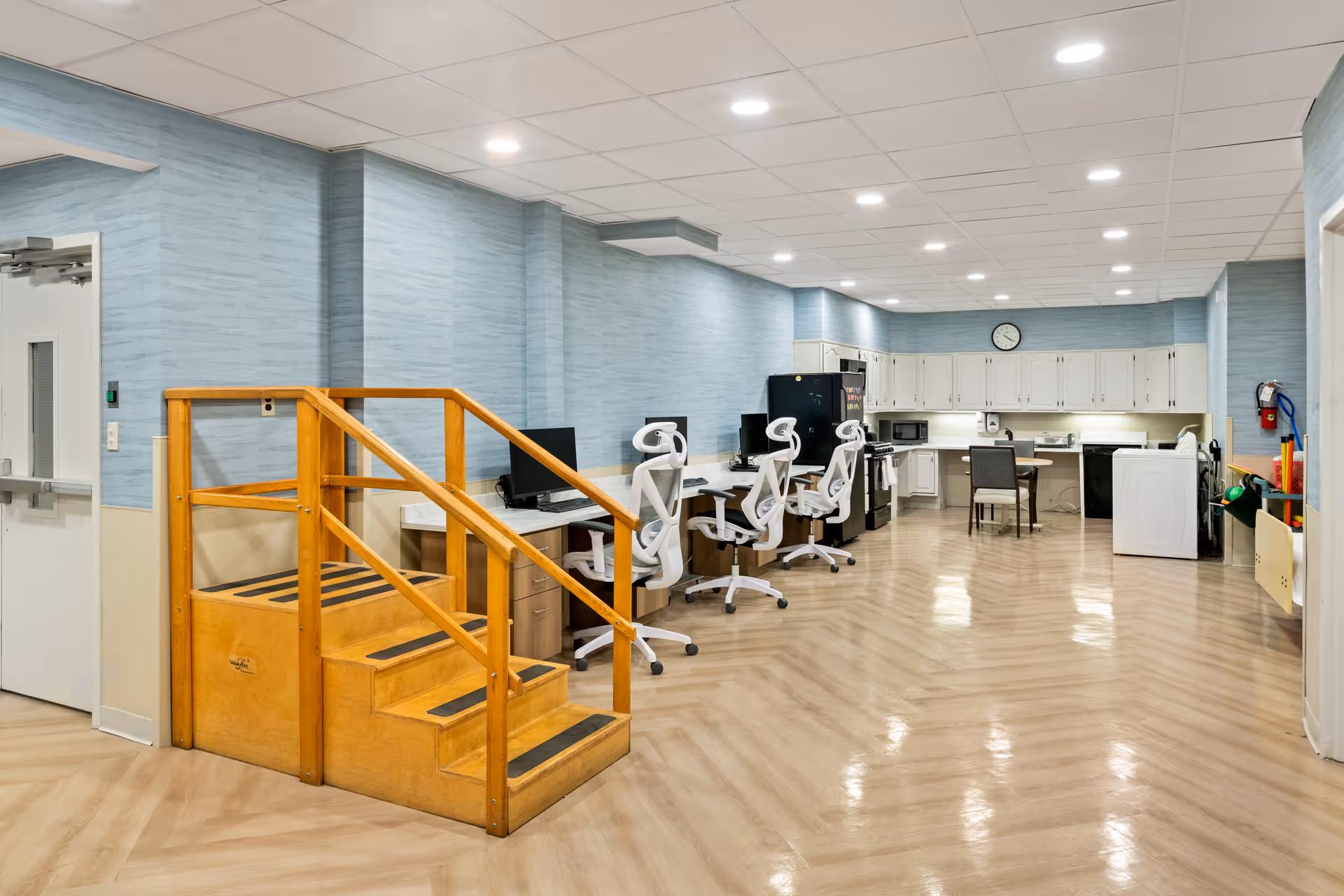 A spacious rehabilitation room with light wood flooring and blue walls. The room features a wooden staircase for physical therapy, several white ergonomic office chairs at desks with computer monitors, and a kitchenette area with white cabinets, a refrigerator, microwave, and a washing machine. A clock is mounted on the wall above the kitchenette.