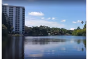 View across a calm lake toward a tall white multi-story building and tree-lined shore under a blue sky with scattered clouds.