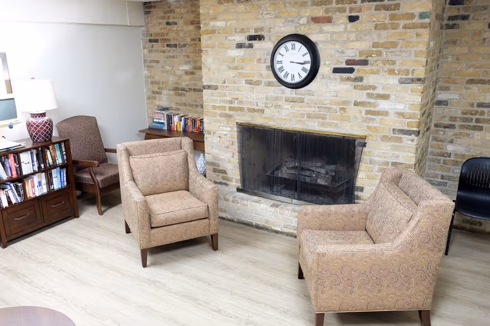 Cozy sitting area with two upholstered armchairs facing a brick fireplace topped by a wall clock and bookshelves to the side.