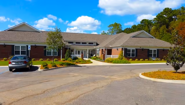 Single-story red-brick memory care facility with a driveway, parking area, landscaping, and blue sky above.