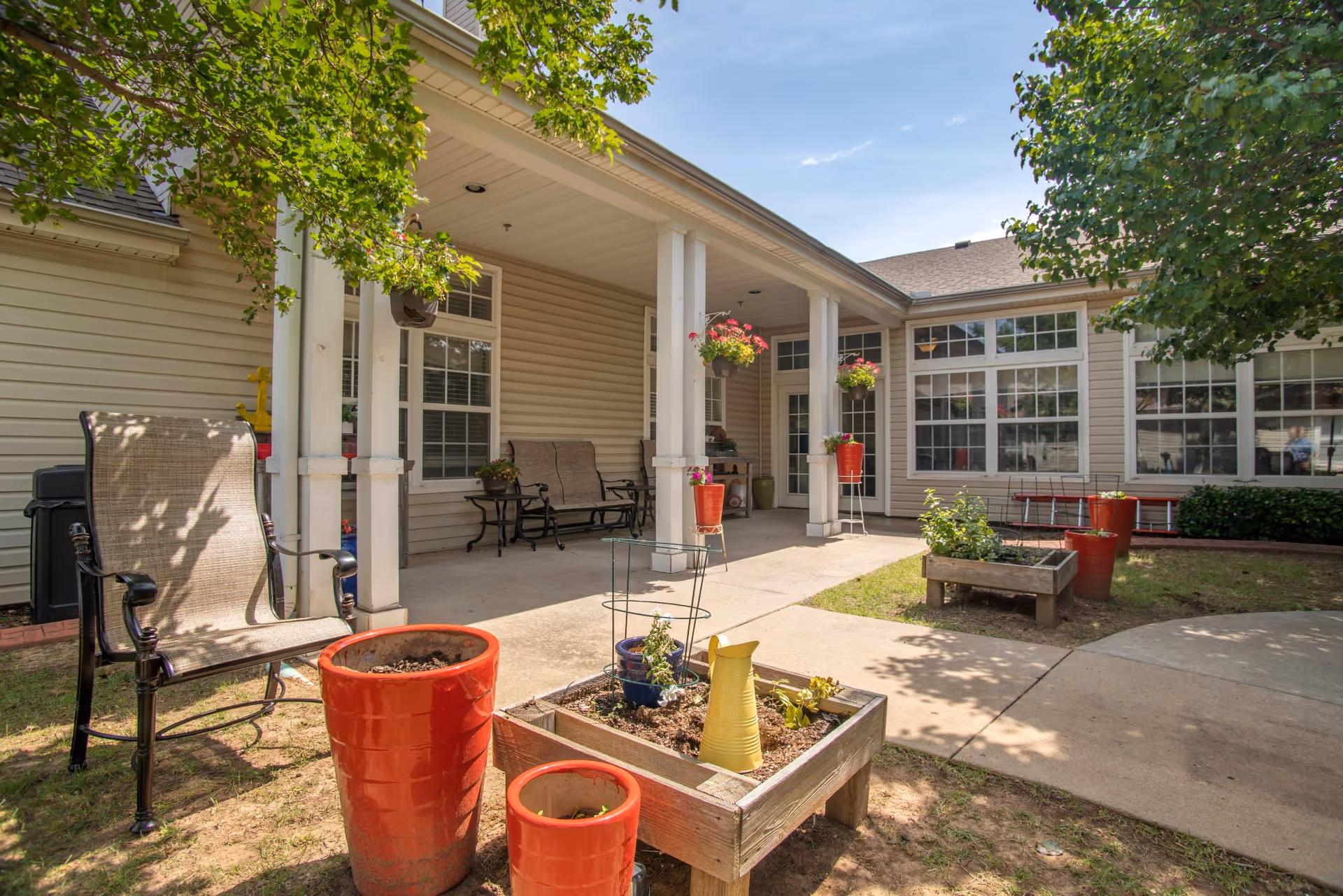 Outdoor patio area at Oxford Springs Tulsa Memory Care with beige siding building, white columns, hanging flower pots, garden planters with plants, and outdoor seating including chairs and benches under a covered porch.