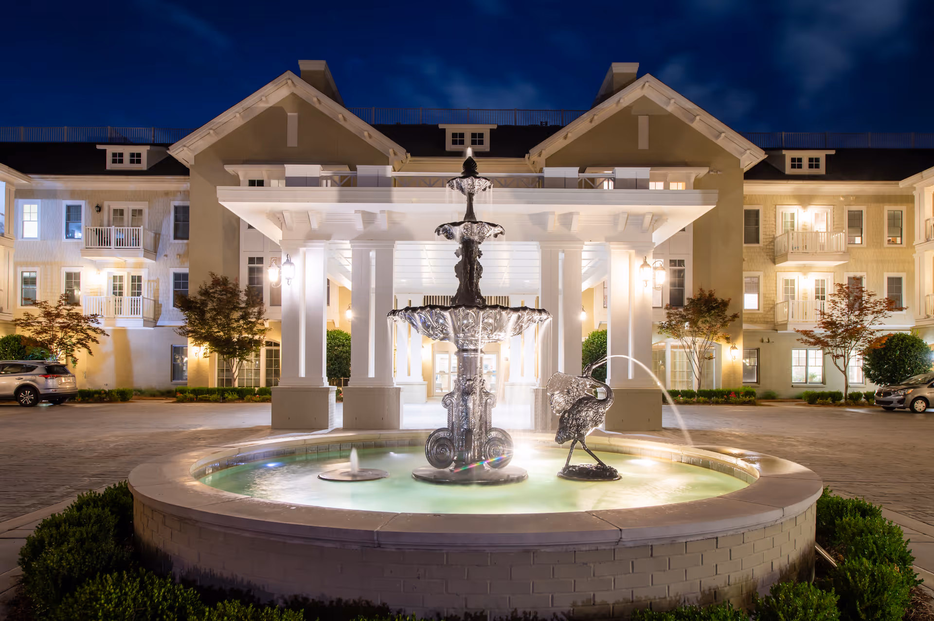 Illuminated entrance of a multi-story senior living building at night with a central fountain in front.