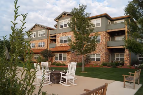 Outdoor patio area with white rocking chairs arranged around a fire pit, surrounded by greenery and trees, in front of a multi-story residential building with stone and green siding.