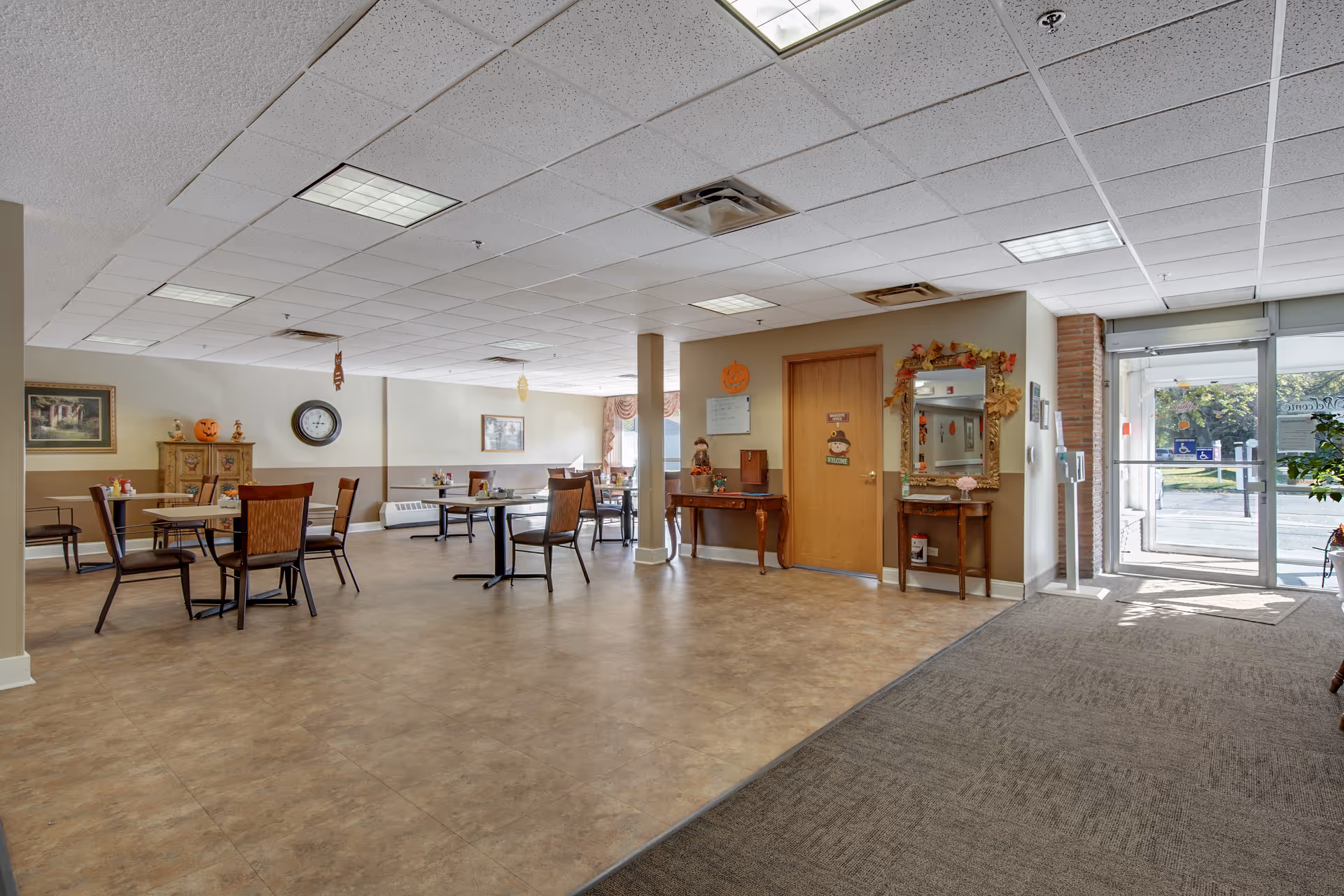 Interior view of a communal dining area in an assisted living facility with several tables and chairs arranged around the room. The space is decorated with autumn-themed items including pumpkins and fall leaves. There is a door and a large mirror on the right wall, and a glass entrance door letting in natural light on the far right.