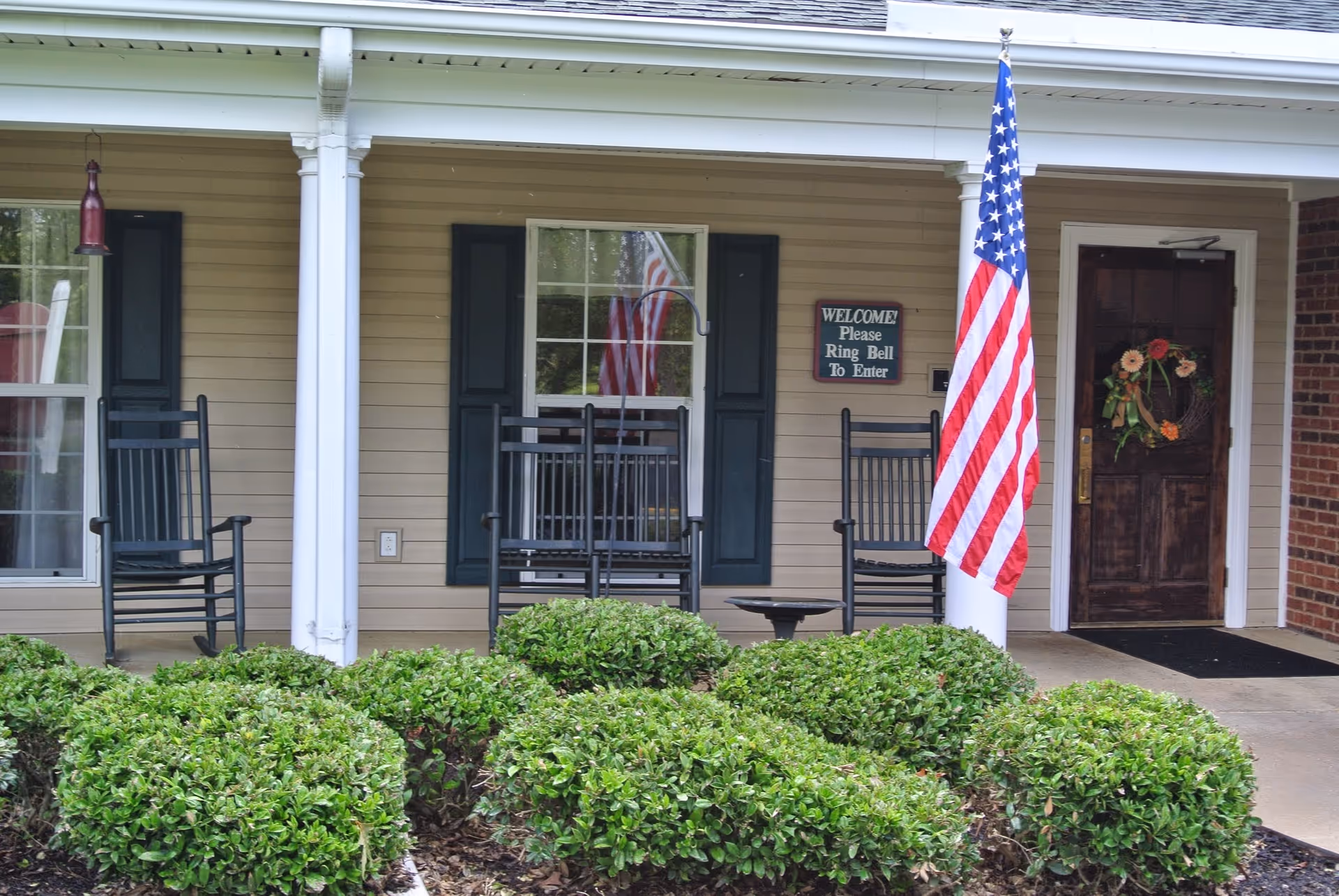 Front porch of a building with beige siding, two white columns, three black rocking chairs, an American flag on a pole, a door with a floral wreath, and a sign that reads 'WELCOME! Please Ring Bell To Enter'. Green bushes are in the foreground.