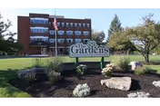 Exterior view of a multi-story brick building with balconies, a flagpole with an American flag, and a landscaped garden area featuring rocks and plants. A sign in front reads 'The Gardens Assisted Living Community'.