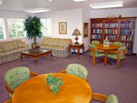 A cozy living room area with two patterned sofas, a wooden coffee table, and a side table with a lamp. There are two round wooden tables with green cushioned chairs around them. A large bookshelf filled with books is against the wall, and a potted plant is near the windows letting in natural light.