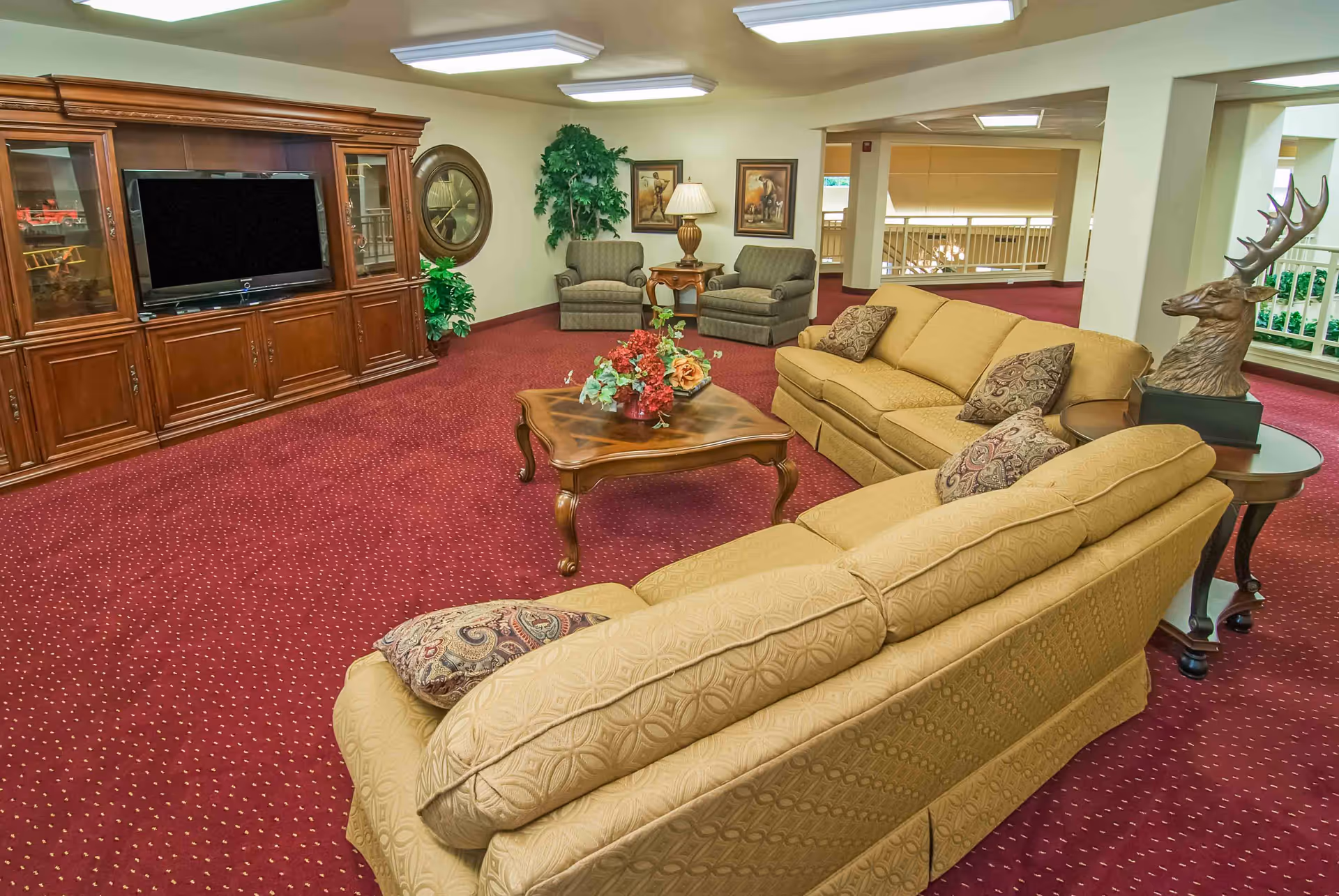 A carpeted communal lounge with beige sofas, armchairs, a coffee table with flowers, and a large wooden entertainment center.