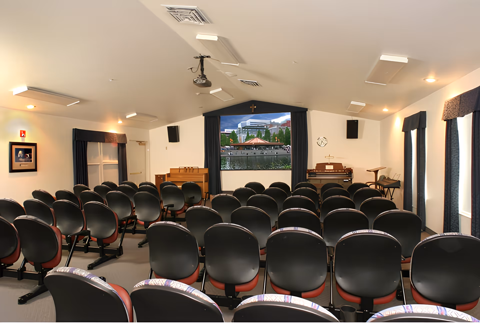 Interior view of a small auditorium or chapel with rows of black and red chairs facing a stage area that has a piano, organ, and a large screen displaying an outdoor scene. The room has white walls, blue curtains on windows, ceiling lights, and speakers mounted on the walls.