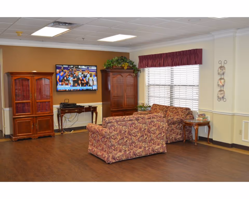 A cozy living room area with two patterned sofas facing a wall-mounted flat-screen TV. The room features wooden cabinets, a small table with board games, a window with blinds and a maroon valance, and decorative plates on the wall. The floor is wooden, and the ceiling has recessed lighting.