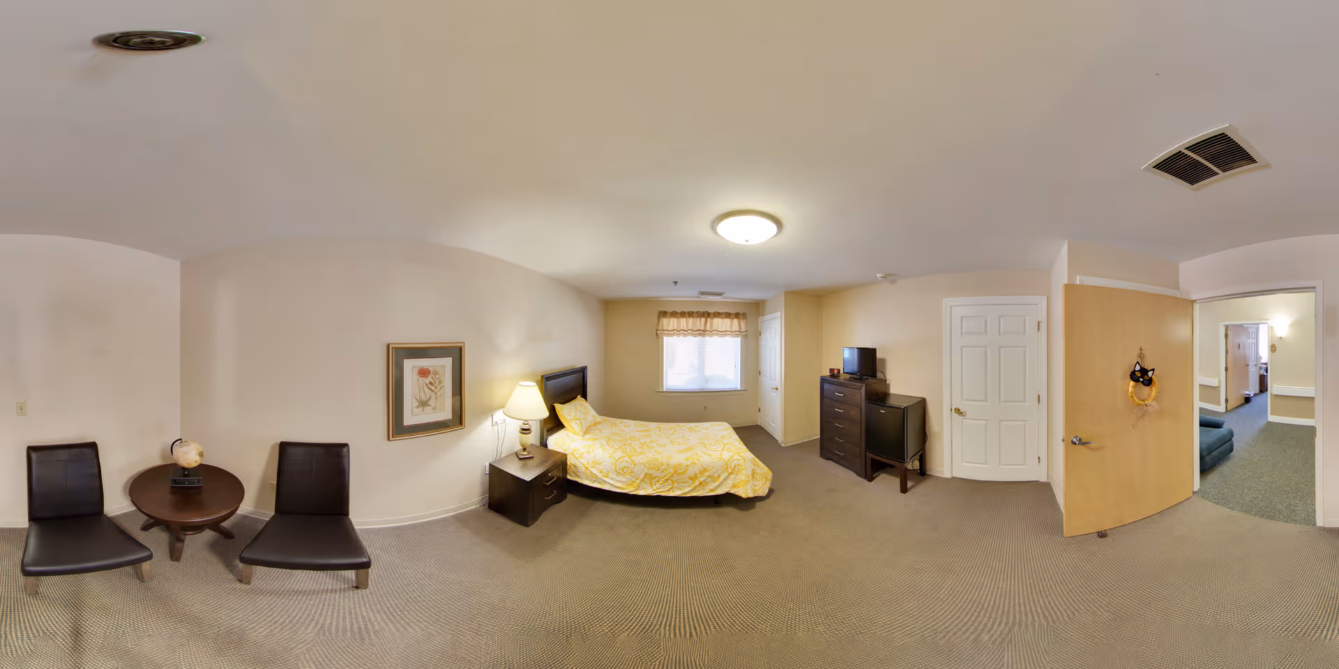 Panoramic bedroom in a senior living facility showing a bed with yellow bedding, nightstand and lamp, dresser with a TV, two chairs with a small table, and an open door to a hallway.